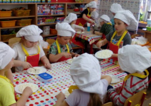 Dzieci przy dwóch stolikach w strojach kucharzy podczas rozgniatania twarogu.